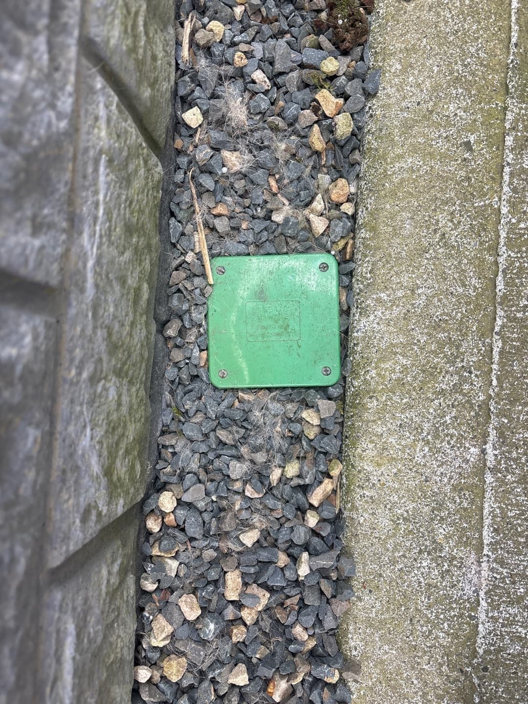 Green inspection lid in gravel beside a wall